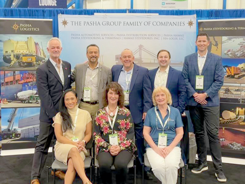 Eight people, dressed in business attire and wearing conference badges, pose and smile in front of a trade show booth for The Pasha Group Family of Companies, featuring maritime and logistics imagery.