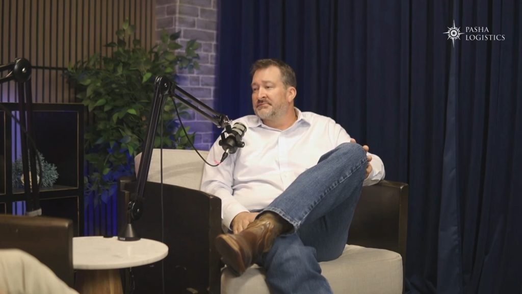 A man in a white shirt and jeans sits with legs crossed in a modern armchair, speaking into a microphone. A small table, plant, and the "Pasha Group" logo are in the background, hinting at a discussion about Pacific Northwest logistics.