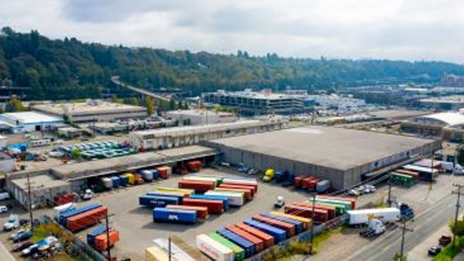 Aerial view of an industrial area in the Pacific Northwest with colorful shipping containers neatly arranged outside Pasha Group warehouse buildings, surrounded by roads, trees, and a forested hillside—showcasing the region’s dynamic growth.