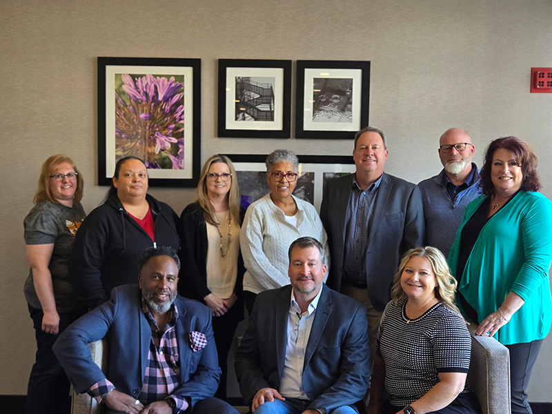 A group of eleven adults, six standing and five sitting, pose together indoors in front of a wall decorated with framed art and photographs. They are smiling and dressed in business casual attire.