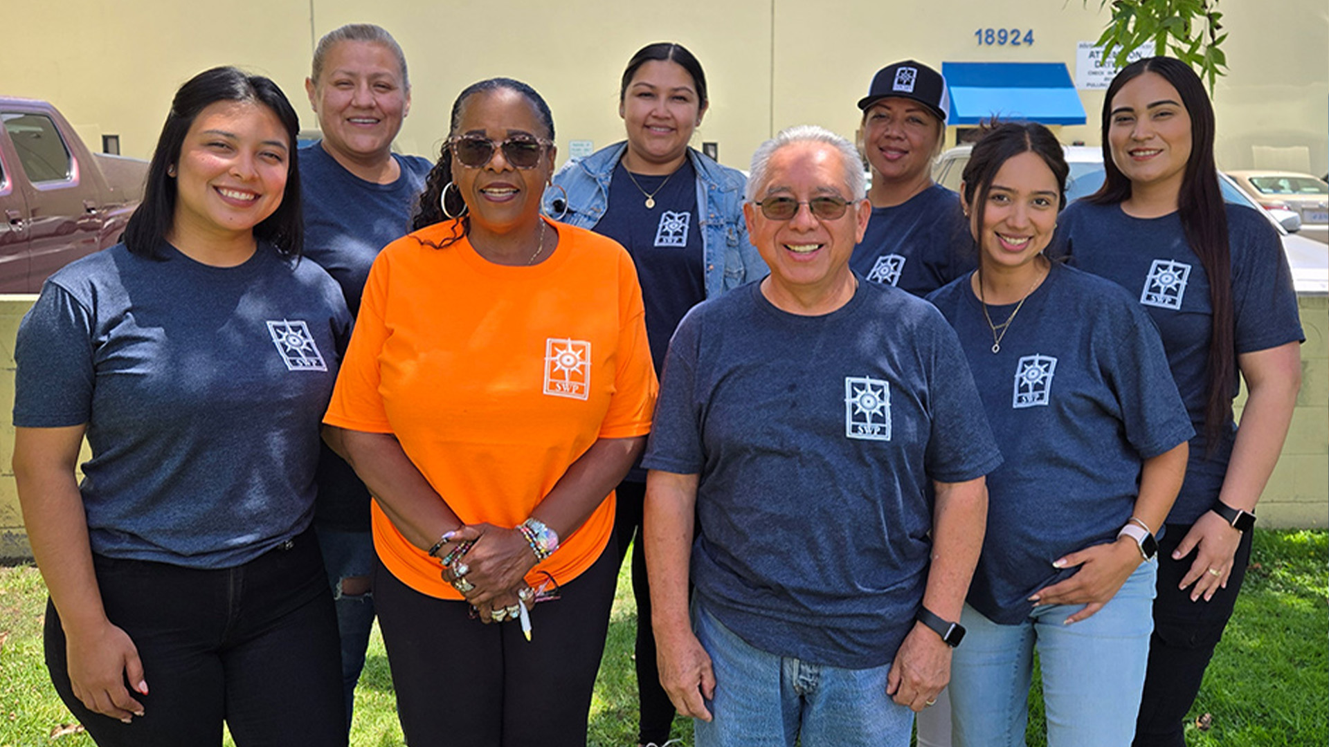 A group of eight smiling people pose outdoors in front of a building. Seven wear matching blue shirts with a white logo; one woman in front wears an orange shirt with the same logo. All appear happy and relaxed.